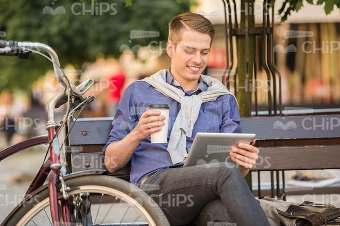 Cheerful Young Man Sitting On Bench, Using Tablet and Holding Coffee Cup Stock Photo