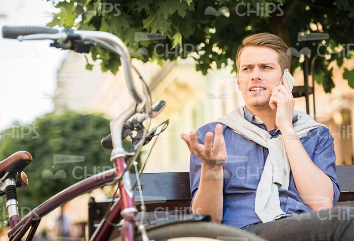 Frowning Man Sitting On Bench While Talking The Phone Stock Photo