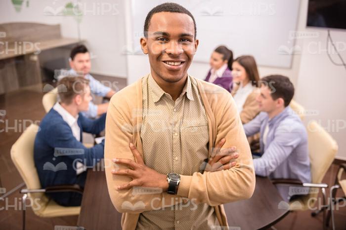 Cheerful Black Young Man Crosses Arms While Stands In Front Of Colleagues Stock Photo
