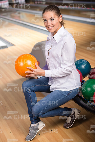 Cheerful Young Woman Sitting Next To Bowling Balls While Holding The Orange One Stock Photo
