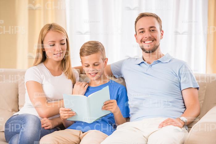 Boy Sitting With Parents And Reading Book Stock Photo