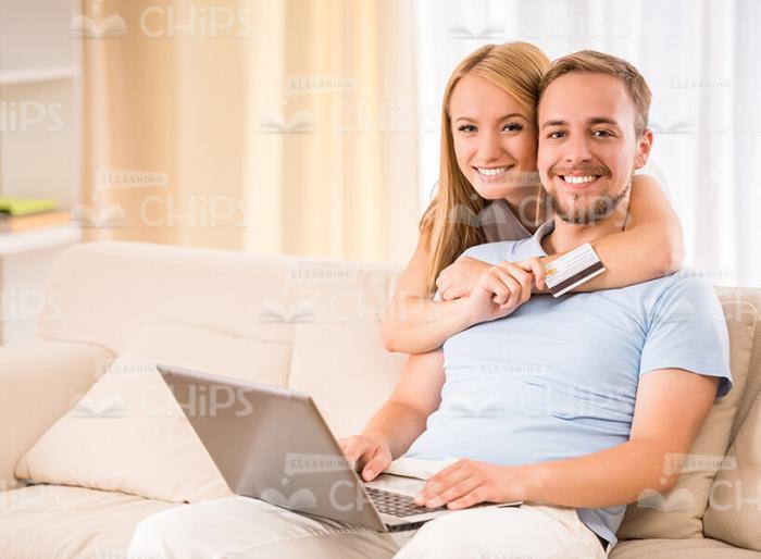 Young Woman Hugs Her Husband Who Sits On Sofa With Laptop Stock Photo
