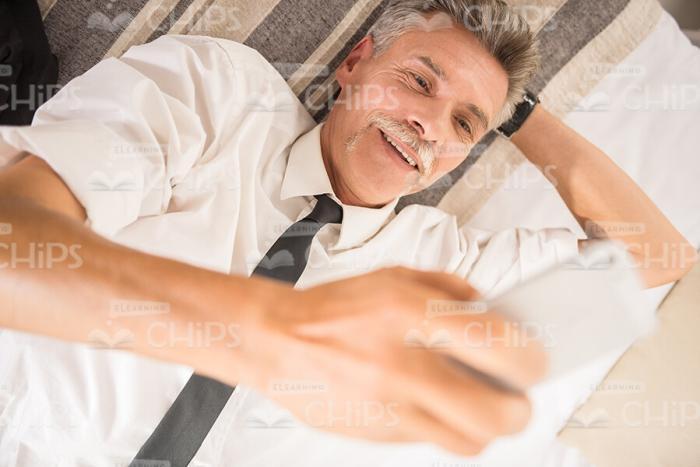 Old Businessman Lying On Bed And Using Phone Stock Photo